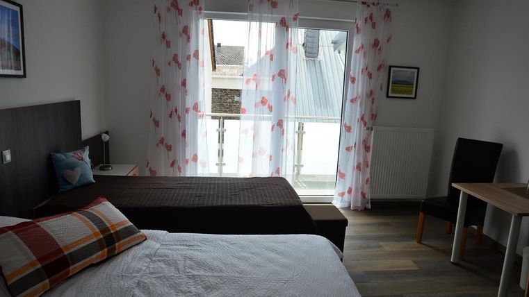 Ein helles Zimmer mit zwei Betten und einem Tisch. Große Fenster mit weißen Vorhängen lassen viel Licht herein.