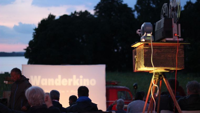 Ein Outdoor-Kinoabend mit Menschen, die einem Film zuschauen. Im Vordergrund steht ein Projektor, der ein Bild auf eine Leinwand projiziert.