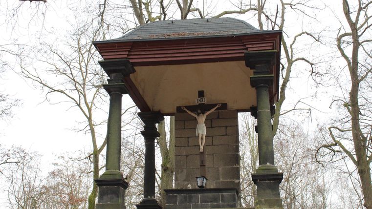 Eine kleine Kapelle umgeben von Bäumen. In der Mitte hängt ein Kreuz mit einer Statue.