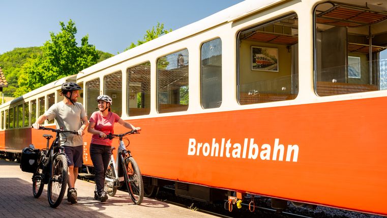 Mann und Frau schieben ihre Fahrräder. Im Hintergrund sieht man einen roten Waggon des Vulkan-Express.