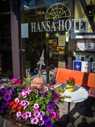 Einladender Eingangsbereich des Hansa Hotels mit einer bunten Blumenbank. Ein Tisch mit sitzenden Stühlen lädt zum Verweilen ein.