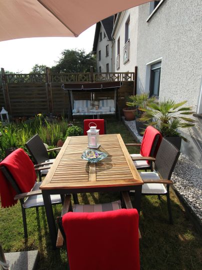 Ein schöner Gartentisch aus Holz mit roten Stühlen. Im Hintergrund sind Pflanzen und ein Haus zu sehen.