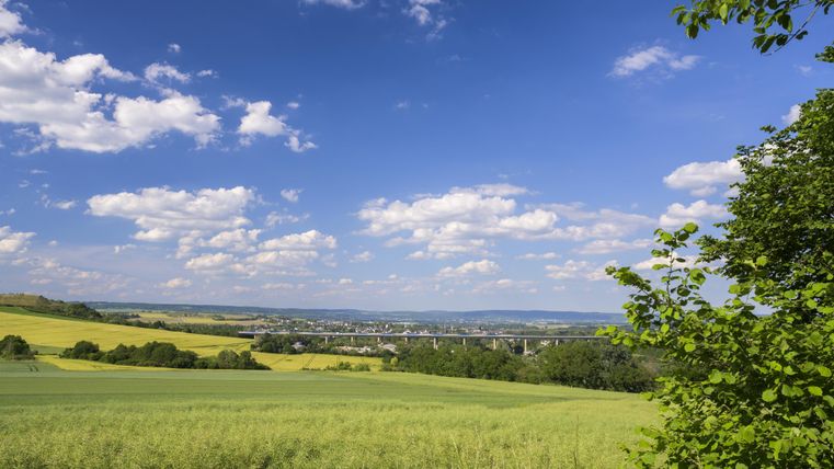 A picturesque landscape with green fields and a clear blue sky. In the background, some clouds and a small village are visible.