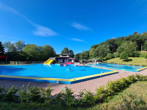 Ein schöner Freibadbereich mit einem blauen Schwimmbecken und einer Rutschenanlage. Umgeben von Bäumen und einer grünen Wiese, strahlt der Ort Ruhe und Erholung aus.