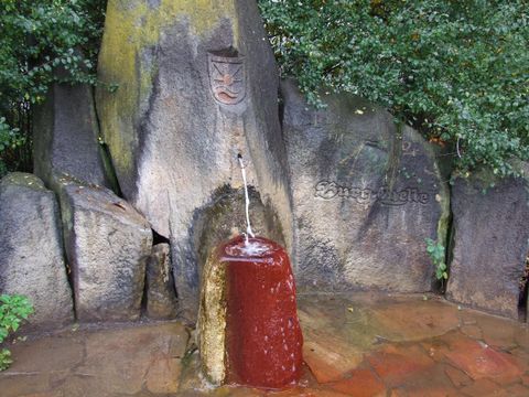 A spring that bubbles out of a rock, surrounded by large stones and greenery. The water has a reddish hue and flows gently from the spring.