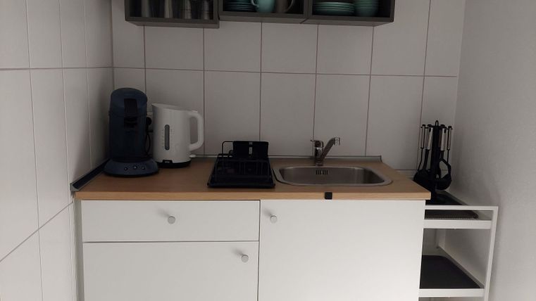 A small, modern kitchenette with white cabinets and a wooden work area. Simple shelves with dishes and kitchen utensils are mounted on the wall.