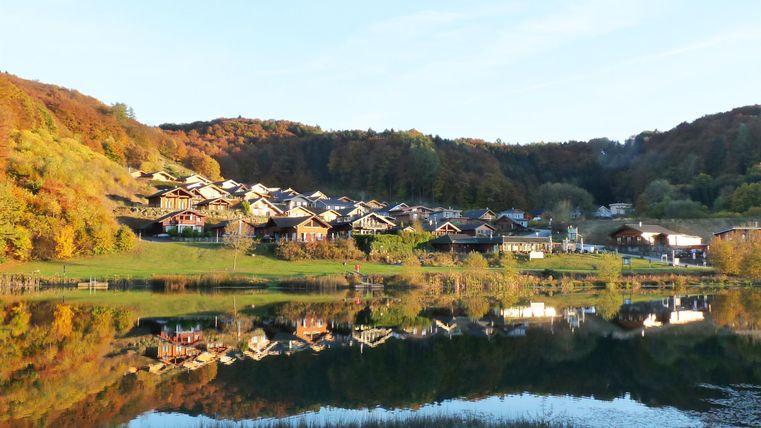 Eine malerische Landschaft mit bunten Bäumen und einem ruhigen See. Die Gebäude spiegeln sich harmonisch im Wasser wider.
