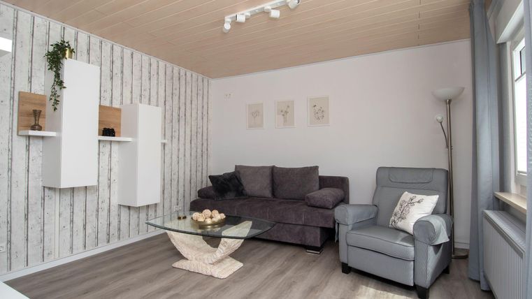 Ein stilvolles Wohnzimmer mit einem grauen Sofa und einem modernen Sessel. An der Wand hängen dekorative Regale und Kunstwerke.