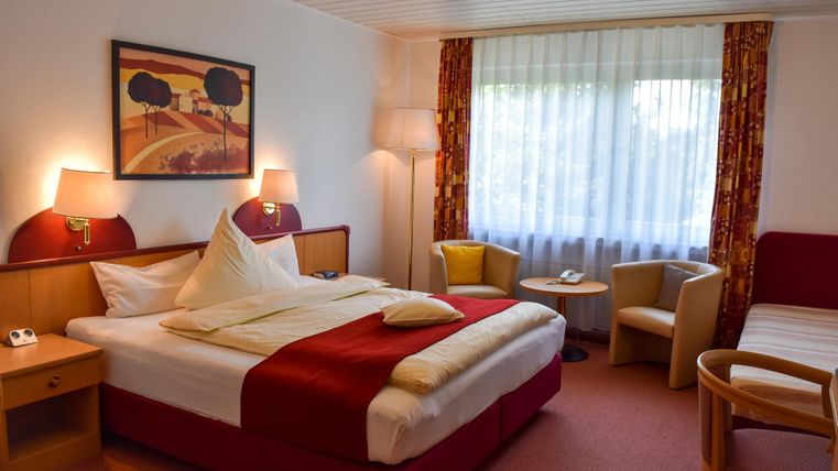 Ein gemütliches Hotelzimmer mit einem großen Bett und bequemen Sesseln. Das Zimmer ist hell und einladend, mit einem Fenster, das Tageslicht hereinlässt.