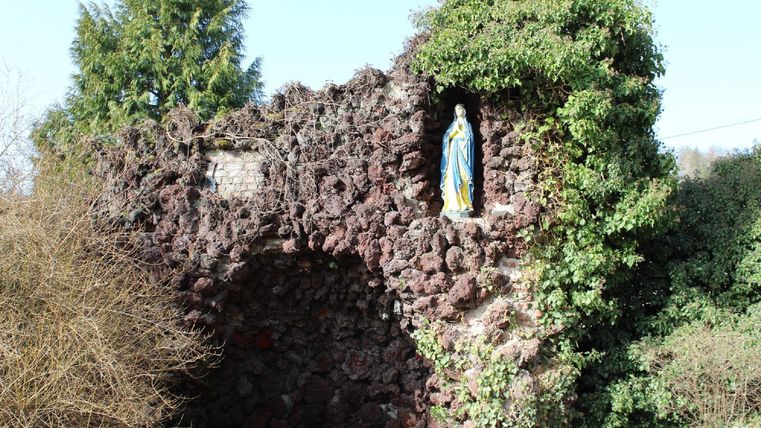 An old, plant-covered wall with a statue of the Virgin Mary. The surroundings are close to nature and somewhat wild.