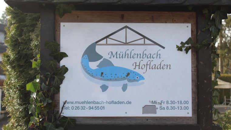 Ein Schild des Mühlenbach Hofladens mit einem blauen Fischsymbol. Es enthält Informationen zu Öffnungszeiten und Kontaktinformationen.