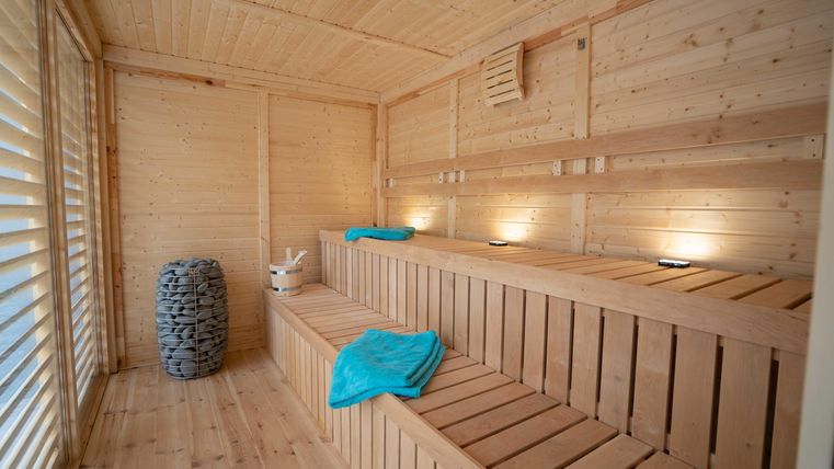 Eine saubere Sauna aus Holz mit gemütlichen Bänken. Auf einer Bank liegen ein hellblaues Handtuch und ein Eimer mit Holzlöffel.