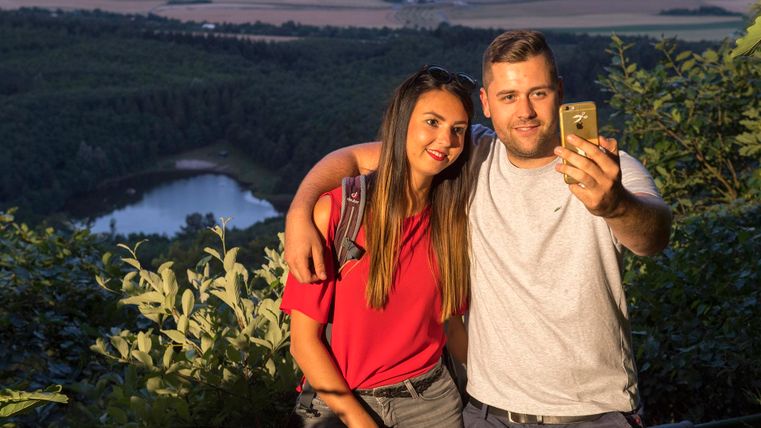 Ein Paar macht ein Selfie vor einer malerischen Aussicht auf das Tal und einen See. Die Sonne scheint und die Umgebung ist grün und einladend.