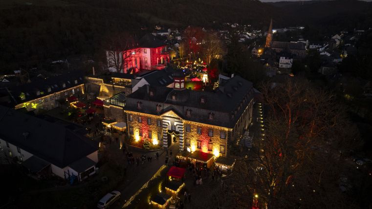Ein historisches Gebäude, das festlich beleuchtet ist. Im Hintergrund ist eine kleine Stadt in der Dämmerung zu sehen.