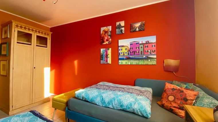 Ein gemütliches Zimmer mit zwei Betten und bunten Wandbildern. Die Wände sind rot gestrichen, und es gibt einen hellen Kleiderschrank sowie eine farbenfrohe Dekoration.