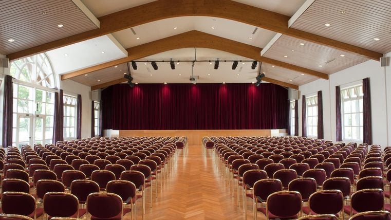 A large event room with many rows of chairs. The room has a wooden floor and large windows that let in a lot of light.