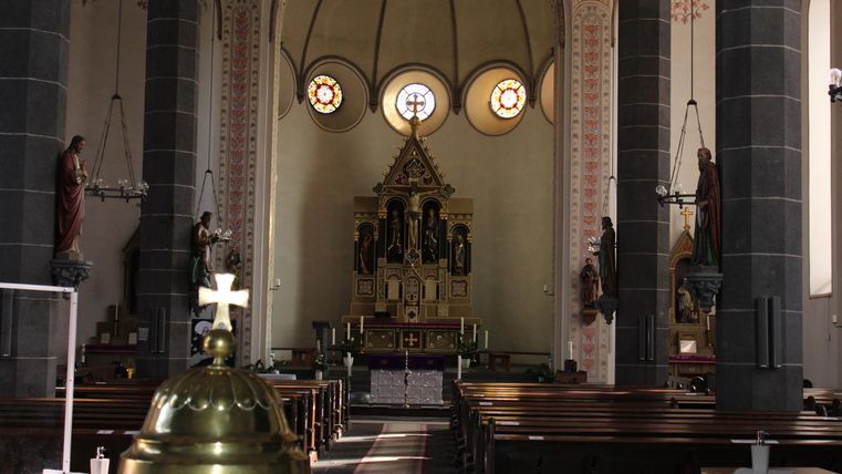 A quiet church with high ceilings and colorful windows. In the background, the altar with religious figures can be seen.