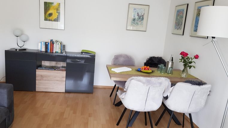Ein modernes Wohnzimmer mit einem Esstisch und eleganten Stühlen. An der Wand hängen Bilder und ein Blumenstrauß steht auf dem Tisch.