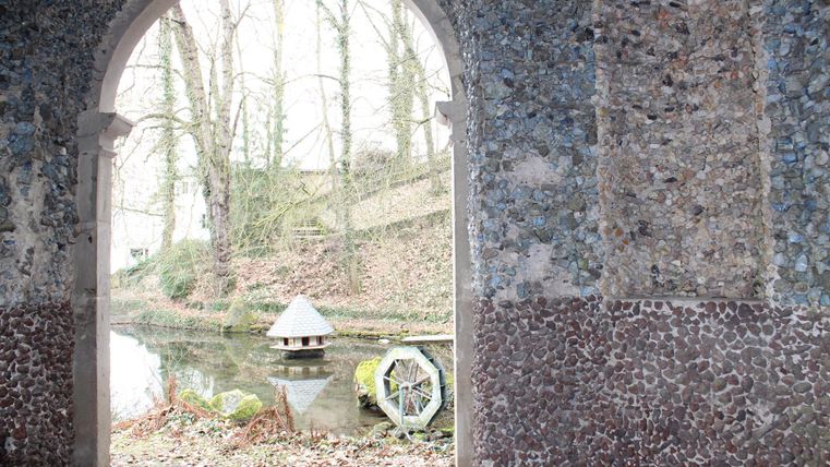 Ein altes Mauerwerk mit einem Torbogen, durch den ein ruhiger Teich zu sehen ist. Im Hintergrund erheben sich Bäume und eine kleine hölzerne Struktur auf dem Wasser.