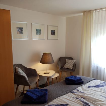 Ein gemütliches Schlafzimmer mit zwei Stühlen und einem Tisch. Die Wände sind mit Bildern dekoriert, und Tageslicht strömt durch die Vorhänge.