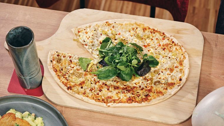 Eine großflächige Pizza mit Käse und frischem Grün auf einem Holzbrett. Daneben befindet sich ein Glas und ein weiterer Teller mit Beilagen.