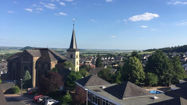Eine ruhige Stadtansicht mit Kirchen und verschiedenen Häusern. Im Hintergrund sind grüne Hügel und ein blauer Himmel zu sehen.