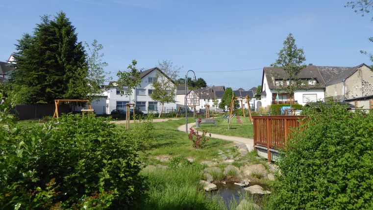Ein schöner Park mit grünen Wiesen und einem kleinen Bach. Im Hintergrund sind Wohnhäuser und Spielgeräte sichtbar.