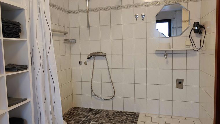 A modern bathroom with a shower and white tiles. On the left side, there is a shelf with towels.