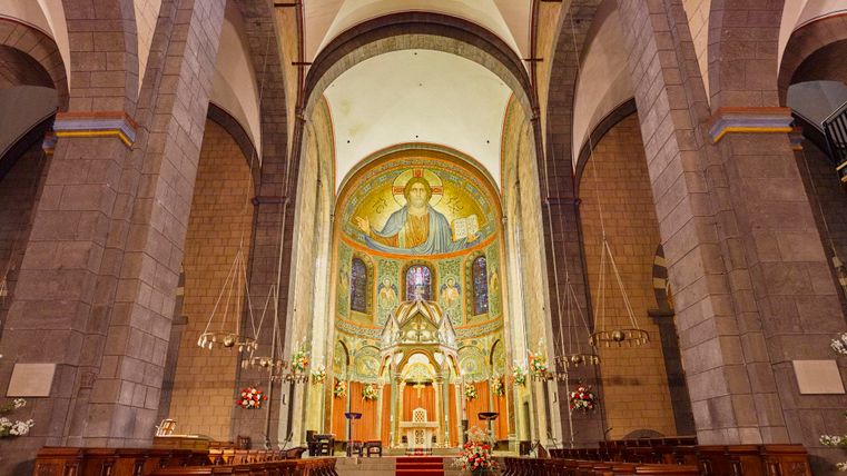 Eine beeindruckende Kirche mit hohen Decken und bunten Wandmalereien. Der rote Teppich führt zum Altar in der Vordergrund.