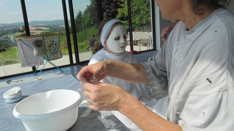 Eine Frau und ein Mädchen sitzen in einem hellen Raum. Das Mädchen hat eine helle Gesichtsmaske auf und die Frau arbeitet an einem Projekt.