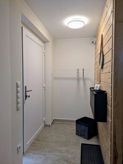 Ein heller Eingangsbereich mit einer weißen Tür und einer Holzvertäfelung an der Wand. Es gibt einen Platz für Jacken und eine schwarze Box auf dem Boden.