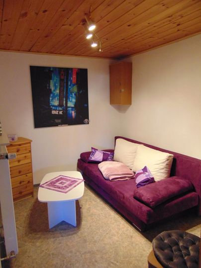A cozy living room with a purple sofa and a low table. A colorful poster hangs on the wall and there are some cushions.