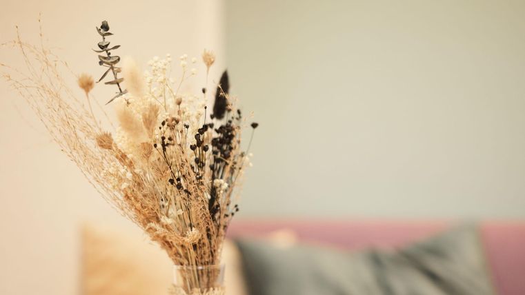 A dried flower arrangement in a vase is in the foreground. The background features a light wall and a soft, color-coordinated pillow.