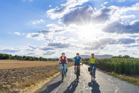 Drei Freunde fahren gemeinsam Fahrrad auf einer sonnigen Straße. Im Hintergrund sind die Felder und der Himmel mit Wolken zu sehen.