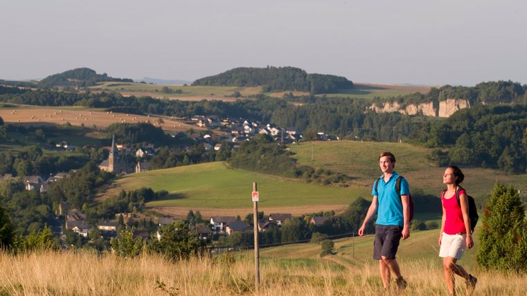 Zwei Personen wandern durch eine hügelige Landschaft mit Dörfern und Feldern im Hintergrund.