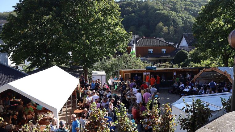 Een drukke buitenruimte met veel mensen en kraampjes. Omringd door bomen en heuvels vindt hier blijkbaar een feest of een evenement plaats.