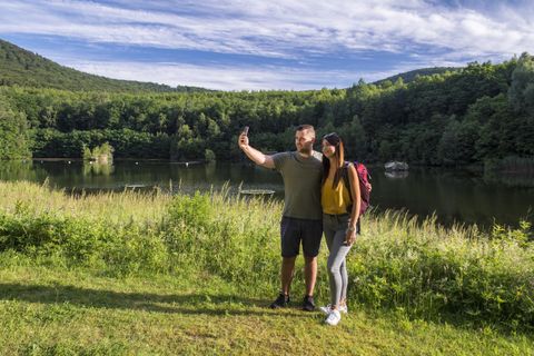 Ein Paar macht ein Selfie am Ufer eines ruhigen Sees, umgeben von grünen Bäumen und sanften Hügeln. Der Himmel ist leicht bewölkt und die Atmosphäre ist entspannt.