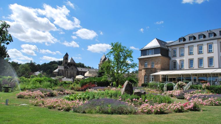 Ein schöner Garten mit bunten Blumen und Grünflächen. Im Hintergrund sind historische Gebäude und ein blauer Himmel zu sehen.