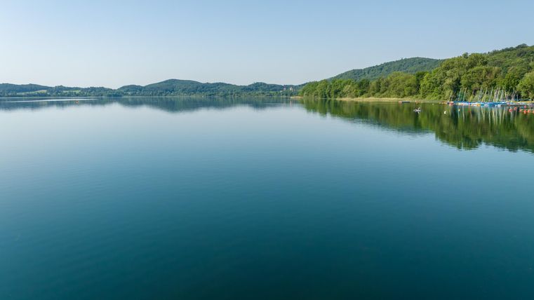 A quiet lake surrounded by gentle hills and dense greenery. The sky is clear and blue, reflecting in the water.