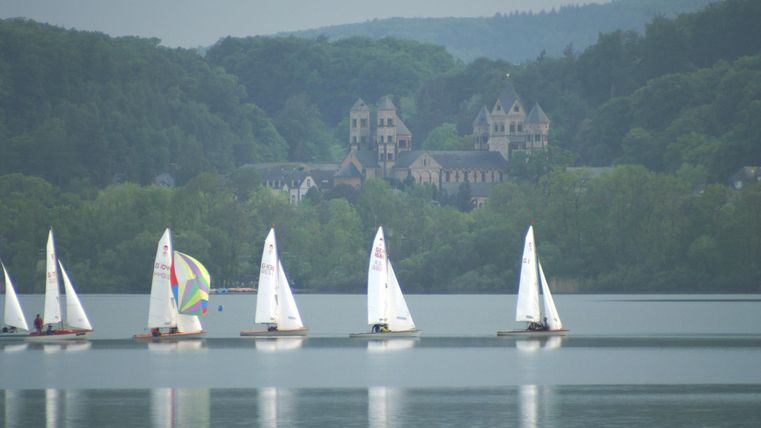 Segelboote fahren ruhig auf einem See, umgeben von grünen Hügeln. Im Hintergrund sind historische Gebäude sichtbar.