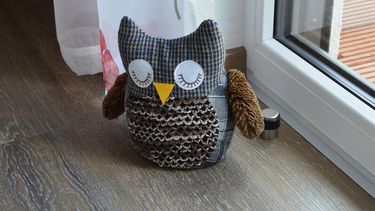 Eine süße Eule aus Stoff mit großen, weißen Augen und einer spitzen Schnauze. Sie steht auf einem Holzboden neben einem Fenster.