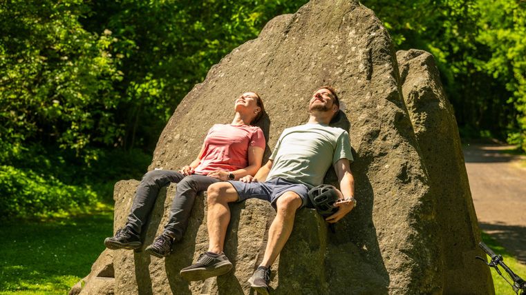 Zwei Personen entspannen auf einem großen Stein im Grünen. Sie genießen die Sonne und die Natur um sie herum.