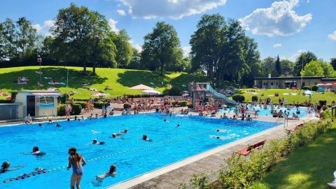 Ein Schwimmbad an einem sonnigen Tag mit vielen Menschen. Umgeben von grünen Wiesen und Bäumen.