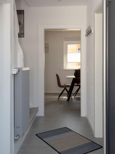 Ein heller Flur mit grauem Boden und einem Läufer. Im Hintergrund sieht man einen Tisch mit einem Stuhl und ein Fenster.