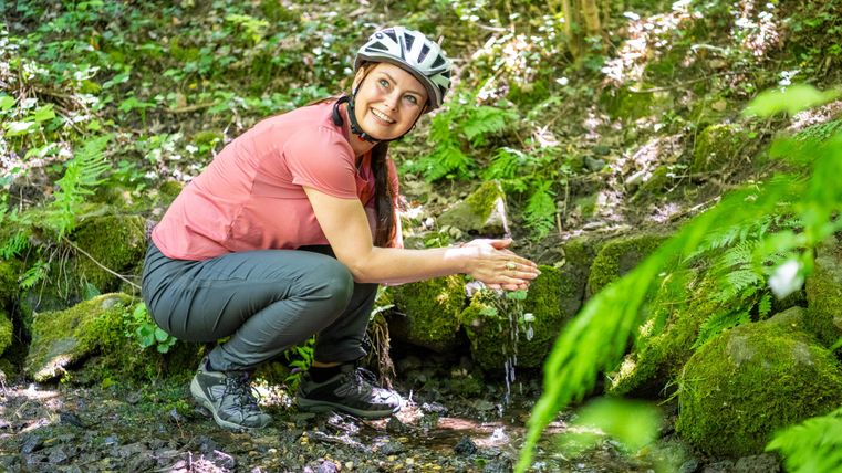 Eine Frau mit einem Fahrradhelm kniet an einem Bach und wäscht sich die Hände. Um sie herum sind grüne Pflanzen und Bäume.