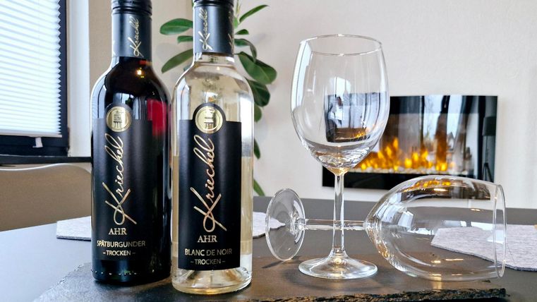 Zwei Flaschen Wein, eine rote und eine weiße, stehen auf einem schwarzen Untersetzer. Daneben steht ein leeres Weinglas und ein umgedrehtes Glas, während im Hintergrund ein warmes Feuer brennt.