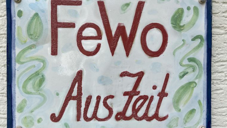 Ein handgemachtes Schild mit der Aufschrift "FeWo AusZeit". Es hat eine bunte, künstlerische Gestaltung.
