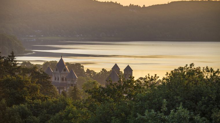 Ein malerischer Blick auf einen See bei Sonnenuntergang, umgeben von sanften Hügeln. Im Vordergrund sind historische Gebäude sichtbar, umgeben von Bäumen.