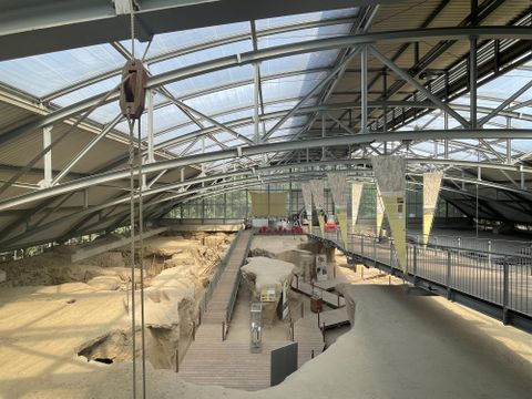 Eine moderne Ausstellungshalle mit einem klaren Glasdach. Im Inneren sind Sandformationen und ein Holzweg für Besucher sichtbar.