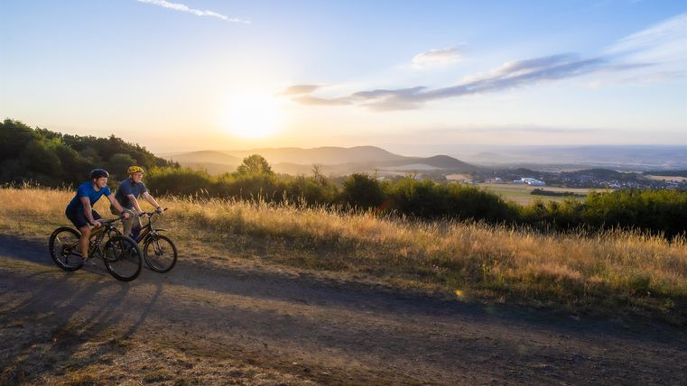 Zwei Radfahrer fahren bei Sonnenuntergang auf einem Feldweg. Im Hintergrund sind sanfte Hügel und ein klarer Himmel zu sehen.